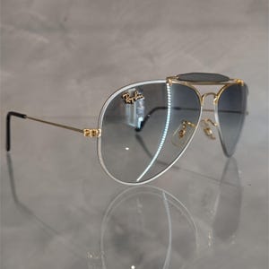 Puede incluir: Gafas de sol aviador con montura dorada, lentes de degradado azul oscuro y barra de cejas negra. Las gafas tienen el logotipo de Ray-Ban en la lente derecha.