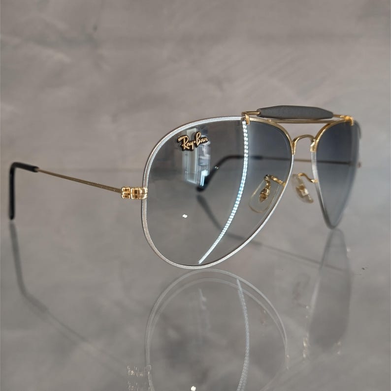 Puede incluir: Gafas de sol aviador con montura dorada, lentes de degradado azul oscuro y barra de cejas negra. Las gafas tienen el logotipo de Ray-Ban en la lente derecha.