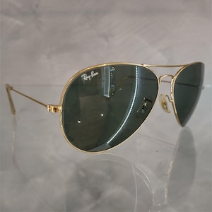 Puede incluir: Gafas de sol aviador doradas con lentes verdes y el logotipo de Ray-Ban en el marco.