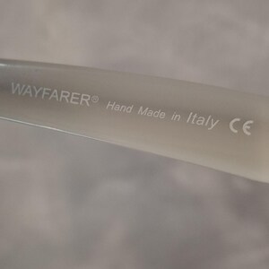 Pu&ograve; includere: Primo piano di un'asta di occhiali Wayfarer di colore chiaro. Il testo "WAYFARER&reg; Hand Made in Italy CE" &egrave; stampato sull'asta. Lo sfondo &egrave; di un colore morbido e neutro.