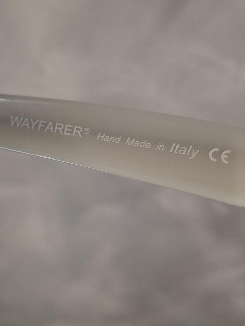 Pu&ograve; includere: Primo piano di un'asta di occhiali Wayfarer di colore chiaro. Il testo "WAYFARER&reg; Hand Made in Italy CE" &egrave; stampato sull'asta. Lo sfondo &egrave; di un colore morbido e neutro.