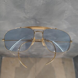 Puede incluir: Gafas de sol aviador doradas con lentes azules y montura cuadrada. Las gafas tienen un puente met&aacute;lico fino y un dise&ntilde;o de doble puente.