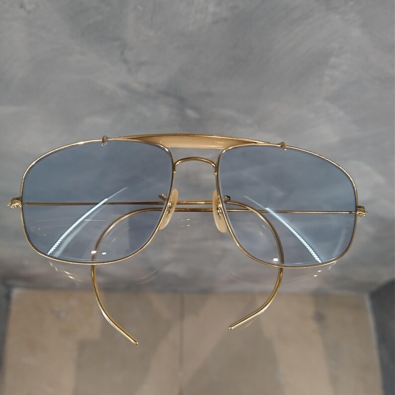 Puede incluir: Gafas de sol aviador doradas con lentes azules y montura cuadrada. Las gafas tienen un puente met&aacute;lico fino y un dise&ntilde;o de doble puente.