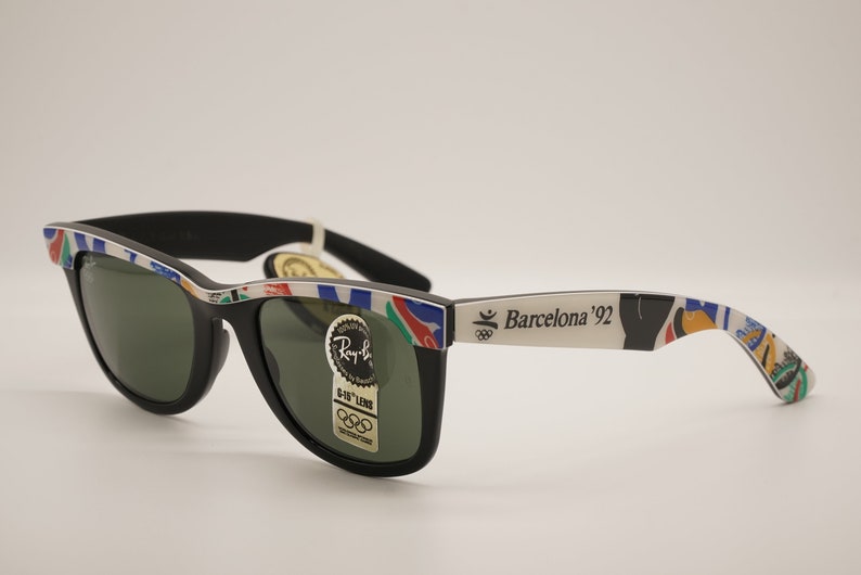 K&ouml;nnte beinhalten: Schwarze Ray-Ban Sonnenbrille mit gr&uuml;nen Gl&auml;sern und einem wei&szlig;en Rahmen mit einem bunten Design und dem Text "Barcelona '92".
