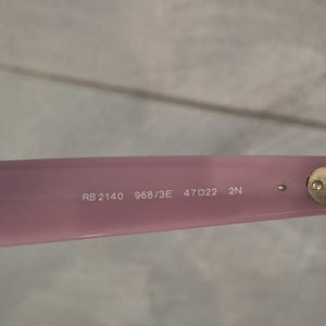 Puede incluir: Primer plano de una montura de gafas rosa claro con el texto "RB 2140 968/3E 47 22 2N" impreso en blanco. La montura tiene una bisagra dorada y un peque&ntilde;o perno redondo.