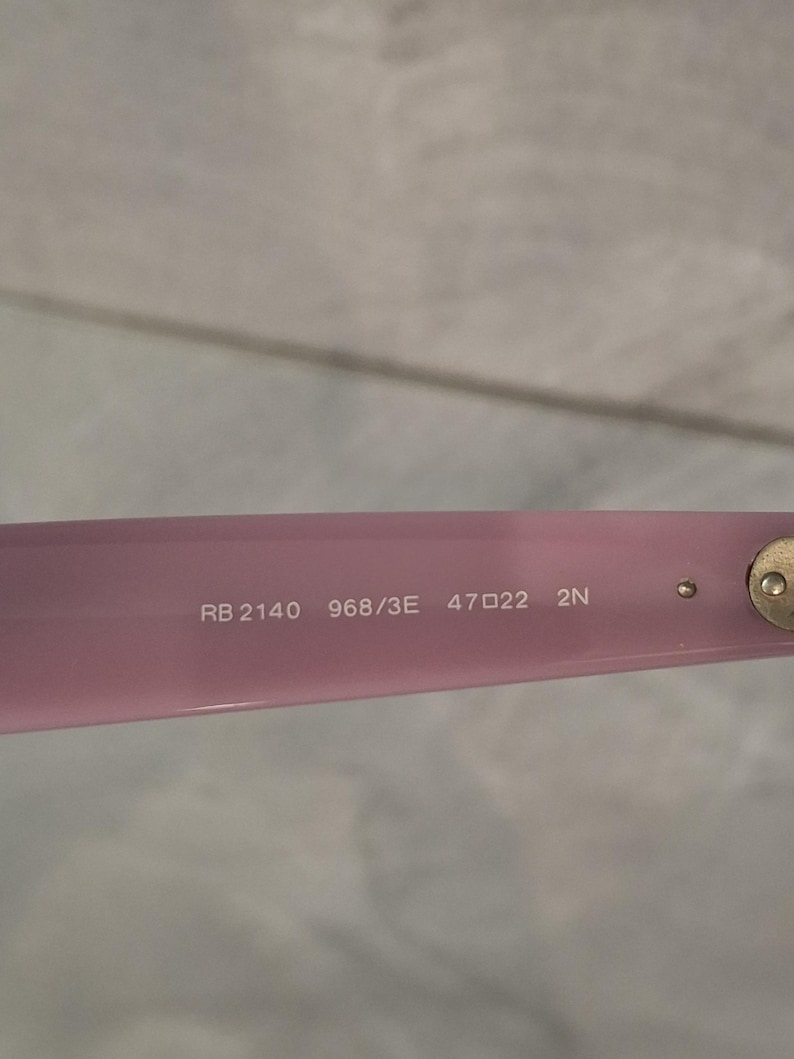 Puede incluir: Primer plano de una montura de gafas rosa claro con el texto "RB 2140 968/3E 47 22 2N" impreso en blanco. La montura tiene una bisagra dorada y un peque&ntilde;o perno redondo.