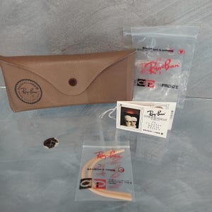 May include: A brown leather Ray-Ban sunglasses case with a Ray-Ban logo and a small white tag with the text "Ray-Ban Ultragradient in Classic Metal Styles" and the text "Bausch & Lomb".