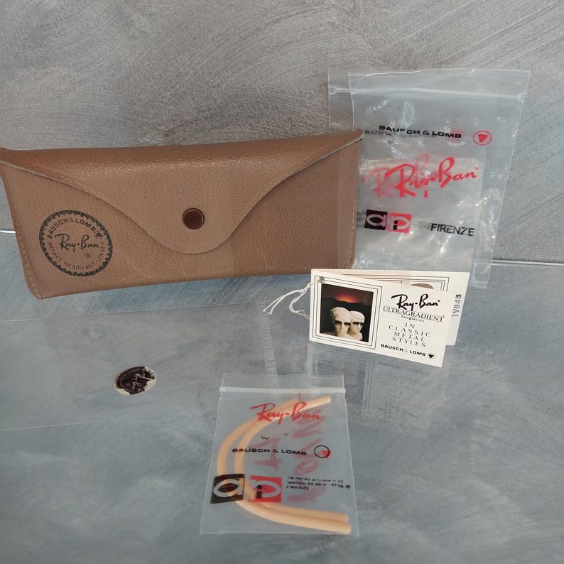 May include: A brown leather Ray-Ban sunglasses case with a Ray-Ban logo and a small white tag with the text "Ray-Ban Ultragradient in Classic Metal Styles" and the text "Bausch & Lomb".