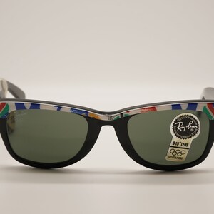 K&ouml;nnte beinhalten: Schwarze Ray-Ban Sonnenbrille mit wei&szlig;em Rahmen und einem bunten olympischen Logo-Design. Die Sonnenbrille hat gr&uuml;ne Gl&auml;ser und einen Aufkleber mit der Aufschrift "G-15 Lens" mit dem olympischen Ringlogo.
