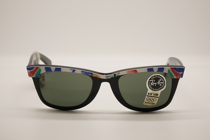 K&ouml;nnte beinhalten: Schwarze Ray-Ban Sonnenbrille mit wei&szlig;em Rahmen und einem bunten olympischen Logo-Design. Die Sonnenbrille hat gr&uuml;ne Gl&auml;ser und einen Aufkleber mit der Aufschrift "G-15 Lens" mit dem olympischen Ringlogo.