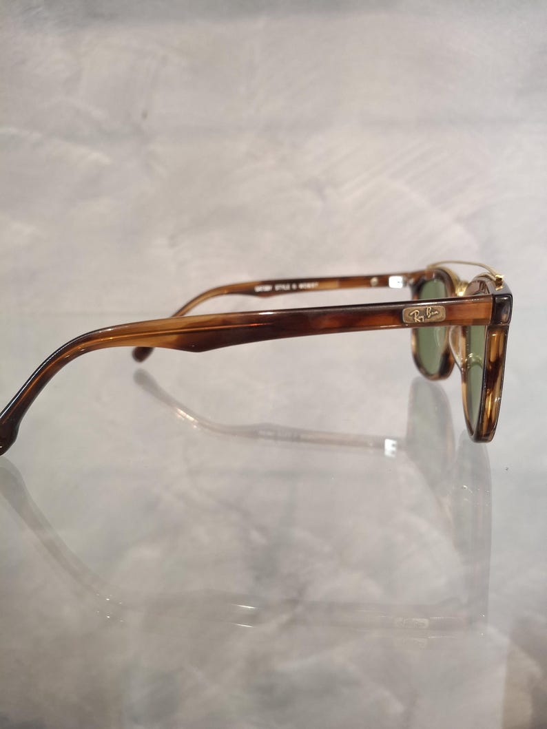 May include: Brown and tortoise shell sunglasses with green lenses. The sunglasses have a classic design with a rectangular frame and a Ray-Ban logo on the temple. The arms are a darker brown color.
