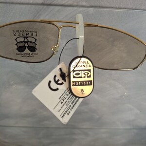 May include: Gold-rimmed sunglasses with tinted lenses. The glasses have a classic aviator style. A white tag with the letters "CE" is attached, along with a black tag that reads "DOPPIA GARANZIA". The lenses are marked "CHANGEABLE LENSES".