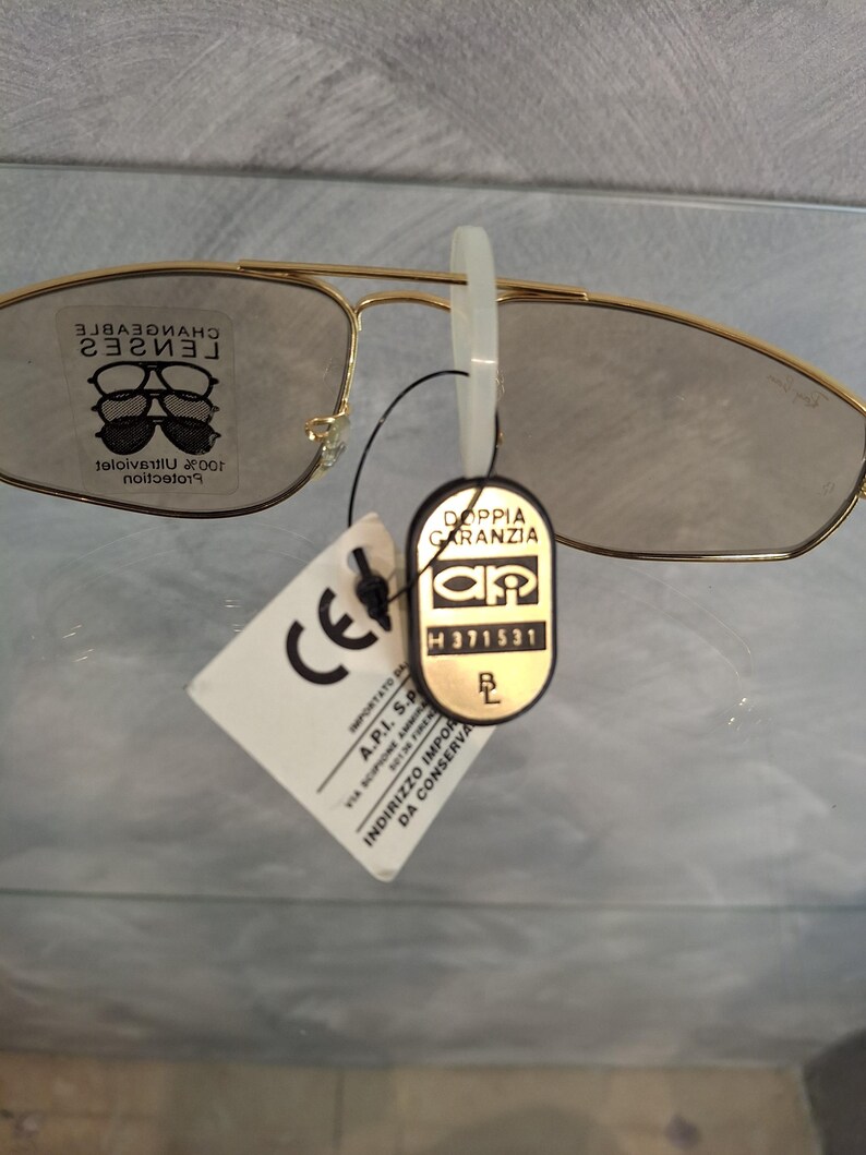 May include: Gold-rimmed sunglasses with tinted lenses. The glasses have a classic aviator style. A white tag with the letters "CE" is attached, along with a black tag that reads "DOPPIA GARANZIA". The lenses are marked "CHANGEABLE LENSES".