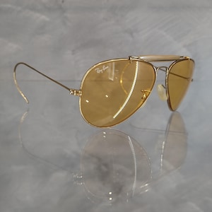 May include: Gold-framed aviator sunglasses with amber-tinted lenses. The Ray-Ban logo is visible on the lens. The sunglasses have a double bridge and curved temples. The lenses are oval-shaped and reflect the light.
