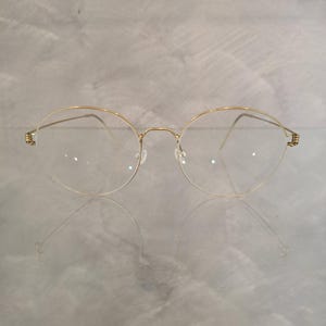 May include: A pair of round, gold-rimmed eyeglasses with clear lenses. The glasses have a delicate, thin frame and are reflected on a light-colored surface. The temples feature a decorative element. The overall design is minimalist and elegant.