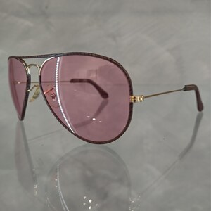 Op de afbeelding: Een paar aviator zonnebrillen met een gouden montuur en roze glazen. De zonnebril heeft een bruine leren rand rond de glazen.