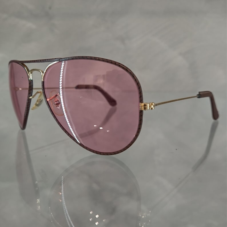Op de afbeelding: Een paar aviator zonnebrillen met een gouden montuur en roze glazen. De zonnebril heeft een bruine leren rand rond de glazen.