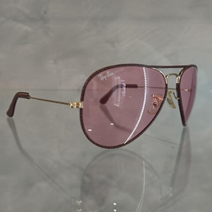 Op de afbeelding: Een paar bruine aviator zonnebrillen met roze glazen. De zonnebril heeft een gouden montuur en het Ray-Ban logo is zichtbaar op het rechter glas.