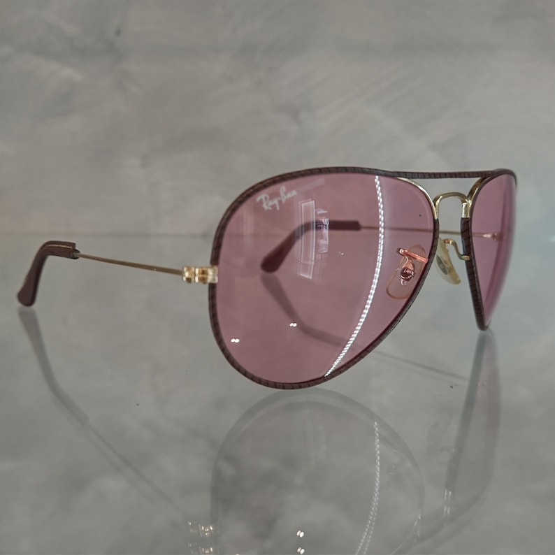 Op de afbeelding: Een paar bruine aviator zonnebrillen met roze glazen. De zonnebril heeft een gouden montuur en het Ray-Ban logo is zichtbaar op het rechter glas.