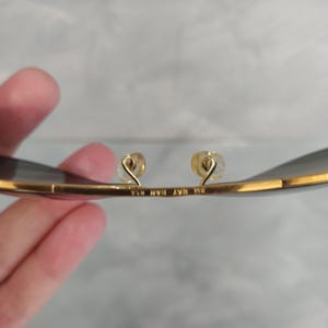 Puede incluir: Primer plano del interior de unas gafas de sol aviador doradas. Las gafas tienen lentes grises oscuras y el texto "BL RAY BAN USA" est&aacute; grabado en la montura de metal.
