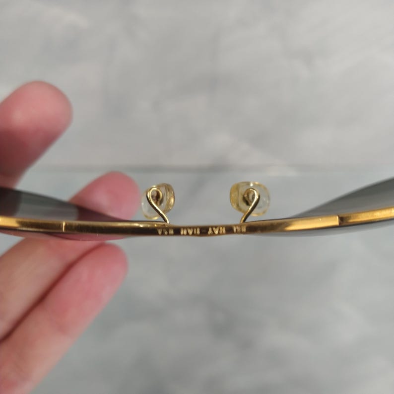 Puede incluir: Primer plano del interior de unas gafas de sol aviador doradas. Las gafas tienen lentes grises oscuras y el texto "BL RAY BAN USA" est&aacute; grabado en la montura de metal.