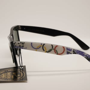 Puede incluir: Gafas de sol negras con una montura blanca que presenta un dise&ntilde;o de anillos ol&iacute;mpicos coloridos. Las gafas de sol tienen una etiqueta negra adjunta con el texto "Producto oficial con licencia de los Juegos Ol&iacute;mpicos de 1984" y el logotipo de los anillos ol&iacute;mpicos.