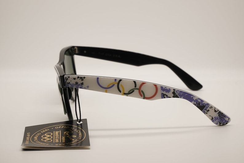 Puede incluir: Gafas de sol negras con una montura blanca que presenta un dise&ntilde;o de anillos ol&iacute;mpicos coloridos. Las gafas de sol tienen una etiqueta negra adjunta con el texto "Producto oficial con licencia de los Juegos Ol&iacute;mpicos de 1984" y el logotipo de los anillos ol&iacute;mpicos.