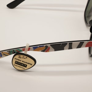 K&ouml;nnte beinhalten: Schwarze Ray-Ban Sonnenbrille mit einem farbenfrohen, abstrakten Druck auf den B&uuml;geln. Die Sonnenbrille hat gr&uuml;ne Gl&auml;ser und einen schwarzen Rahmen. Ein kleines, schwarzes Etikett mit dem Text "Ray-Ban Double Guarantee D074078 Spanien-Portugal" ist am B&uuml;gel befestigt.