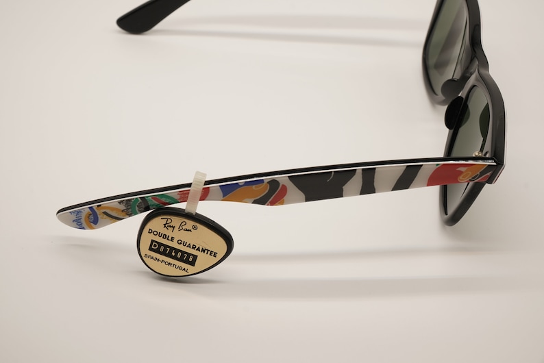 K&ouml;nnte beinhalten: Schwarze Ray-Ban Sonnenbrille mit einem farbenfrohen, abstrakten Druck auf den B&uuml;geln. Die Sonnenbrille hat gr&uuml;ne Gl&auml;ser und einen schwarzen Rahmen. Ein kleines, schwarzes Etikett mit dem Text "Ray-Ban Double Guarantee D074078 Spanien-Portugal" ist am B&uuml;gel befestigt.