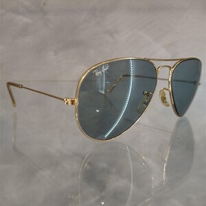 May include: Gold-framed aviator sunglasses with blue-tinted lenses. The Ray-Ban logo is visible on the upper left lens. The sunglasses have a double bridge and a classic teardrop shape.