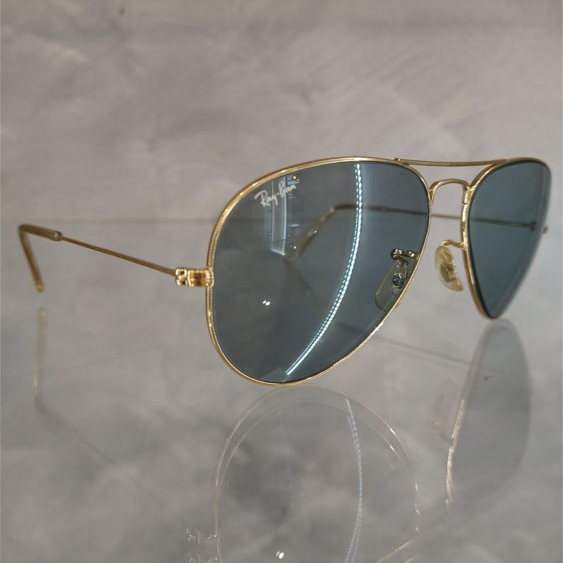 May include: Gold-framed aviator sunglasses with blue-tinted lenses. The Ray-Ban logo is visible on the upper left lens. The sunglasses have a double bridge and a classic teardrop shape.