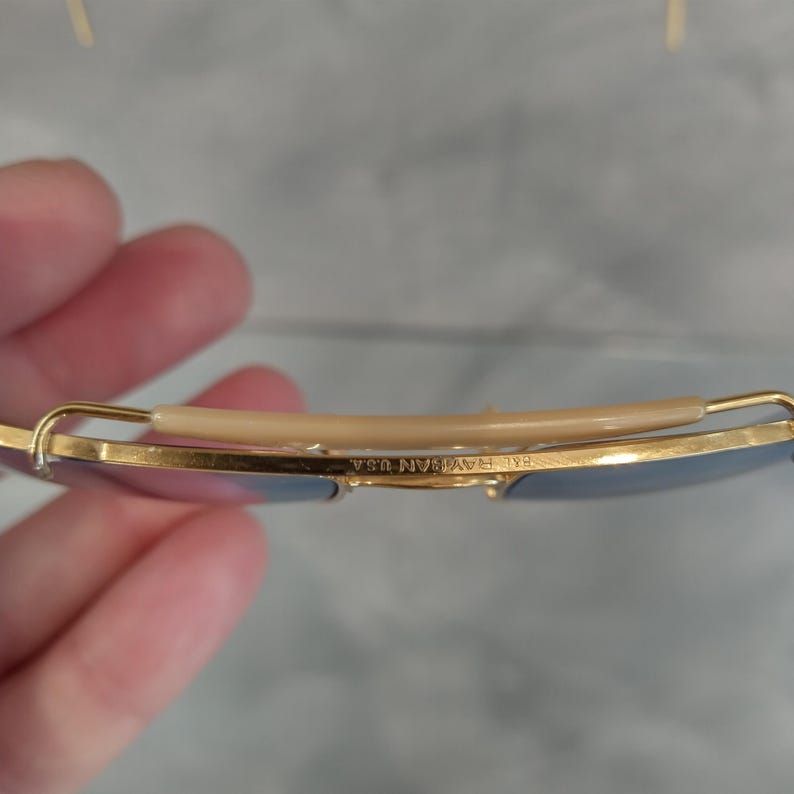 Puede incluir: Primer plano del marco de metal dorado de unas gafas de sol. El marco tiene un puente nasal de pl&aacute;stico marr&oacute;n y el texto "B&L RAY-BAN USA" est&aacute; grabado en el marco.