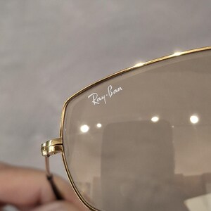 May include: Close-up of Ray-Ban sunglasses with a gold frame and brown lenses. The Ray-Ban logo is visible on the upper left lens. The sunglasses have a classic, rectangular shape.