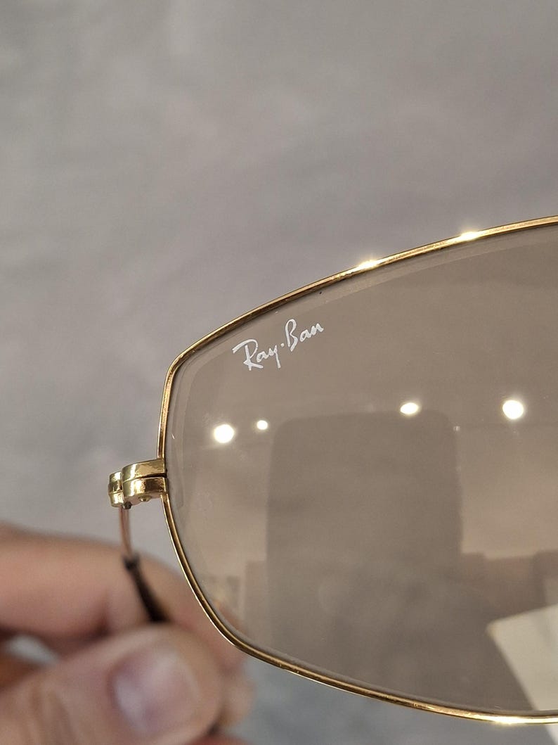 May include: Close-up of Ray-Ban sunglasses with a gold frame and brown lenses. The Ray-Ban logo is visible on the upper left lens. The sunglasses have a classic, rectangular shape.