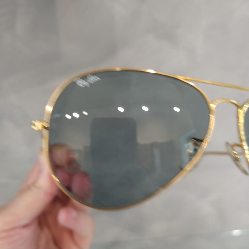 Puede incluir: Gafas de sol aviador con montura dorada y lentes grises oscuros. Las gafas se sostienen en una mano.