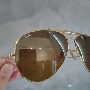 May include: Gold-tone aviator sunglasses with brown mirrored lenses. The sunglasses have a Ray-Ban logo on the left lens.