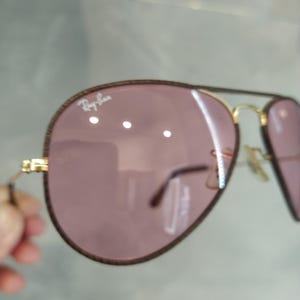 Op de afbeelding: Een paar aviator zonnebrillen met een gouden montuur en roze glazen. De zonnebril heeft een bruine leren rand rond de glazen en het Ray-Ban logo is zichtbaar op het rechter glas.