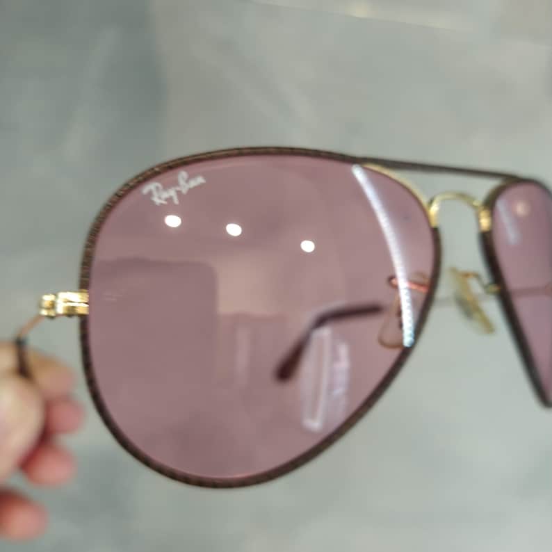 Op de afbeelding: Een paar aviator zonnebrillen met een gouden montuur en roze glazen. De zonnebril heeft een bruine leren rand rond de glazen en het Ray-Ban logo is zichtbaar op het rechter glas.