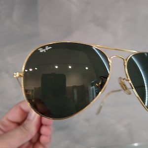 Puede incluir: Gafas de sol aviador con montura dorada y lentes verde oscuro. Las gafas tienen el logotipo de Ray-Ban en la lente izquierda.