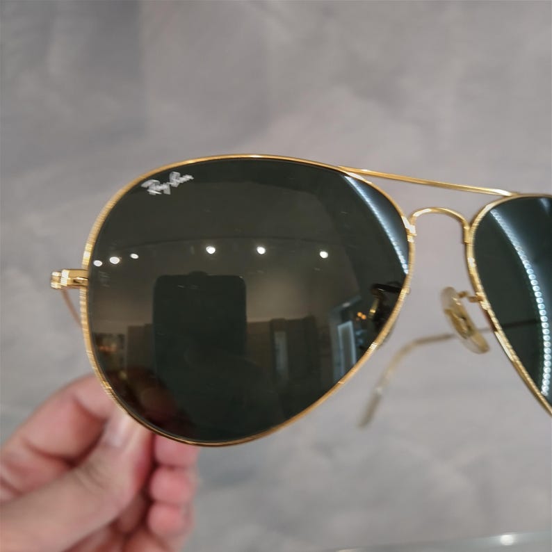 Puede incluir: Gafas de sol aviador con montura dorada y lentes verde oscuro. Las gafas tienen el logotipo de Ray-Ban en la lente izquierda.