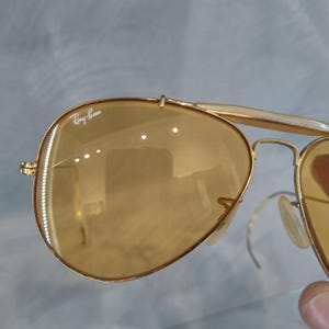 May include: Gold-framed Ray-Ban sunglasses with amber-tinted lenses. The sunglasses have a classic aviator design with a double bridge and nose pads. The Ray-Ban logo is visible on the top left lens.