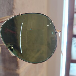 May include: Close-up of a pair of aviator sunglasses with green lenses. The sunglasses have a gold metal frame and the lenses have a subtle green tint. The sunglasses are reflecting light, creating a glare on the lenses.