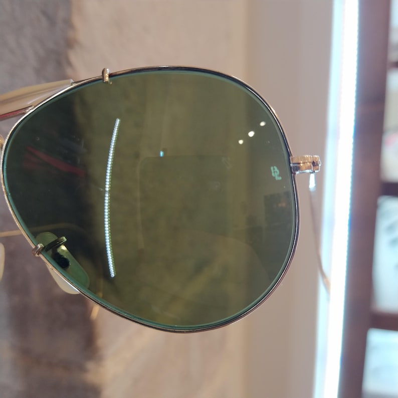 May include: Close-up of a pair of aviator sunglasses with green lenses. The sunglasses have a gold metal frame and the lenses have a subtle green tint. The sunglasses are reflecting light, creating a glare on the lenses.