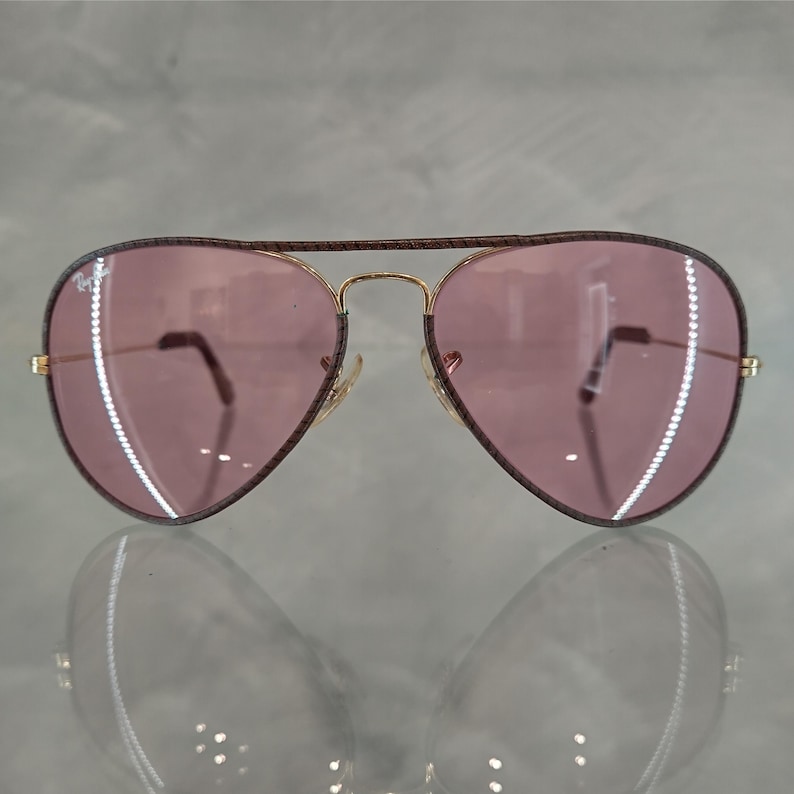 Op de afbeelding: Een paar aviator zonnebrillen met gouden monturen en roze glazen. De monturen hebben een bruin, getextureerd patroon.