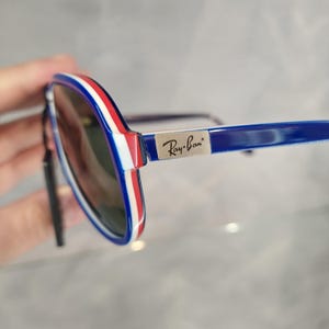 May include: Blue and white aviator sunglasses with red stripes on the arms. The sunglasses have a silver Ray-Ban logo on the right arm.