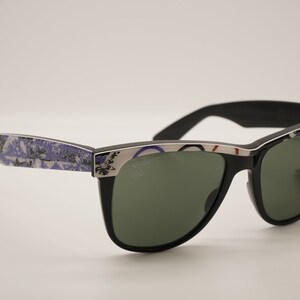 Puede incluir: Gafas de sol negras con lentes verdes y un marco estampado en azul y blanco. Las gafas tienen un borde plateado y el logotipo de Ray-Ban es visible en la lente derecha.