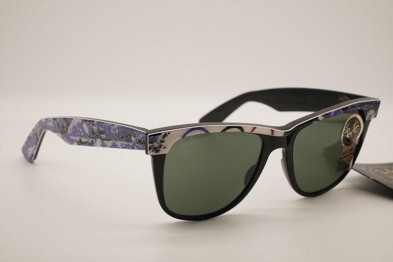 Puede incluir: Gafas de sol negras con lentes verdes y un marco estampado en azul y blanco. Las gafas tienen un borde plateado y el logotipo de Ray-Ban es visible en la lente derecha.