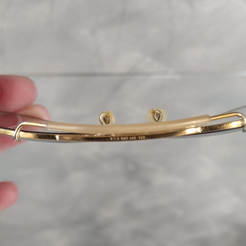 May include: Close-up of the underside of a pair of gold-tone glasses. The glasses have a thin metal frame and clear lenses. The inscription "W.G. & Co. 1/10 12K GF" is visible on the frame.