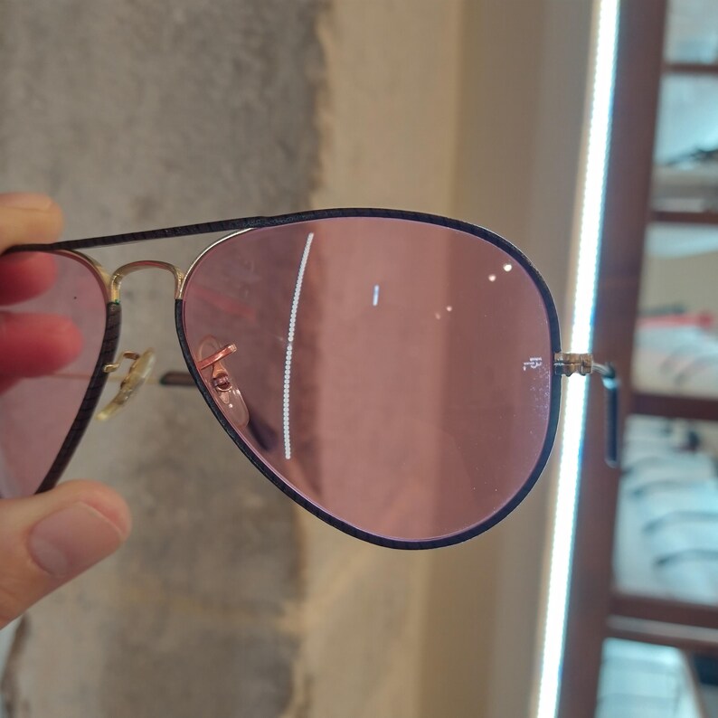 Op de afbeelding: Een paar aviator zonnebrillen met een zwart montuur en roze glazen. De zonnebril wordt in de hand gehouden.