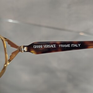 Pu&ograve; includere: Primo piano della montatura marrone e dorata di un paio di occhiali. La montatura ha la scritta "GIANNI VERSACE FRAME ITALY" stampata su di essa.